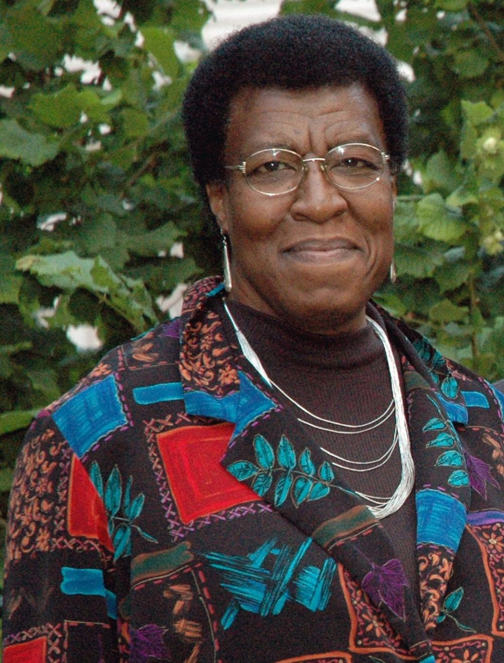 A portrait of author Octavia Butler, photographed outside against a green background. She smiles slightly, and has a short hairstyle, gold wire glasses, and a colorful shirt. 