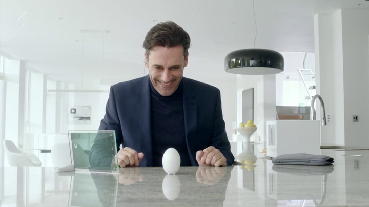 A man sits at a kitchen counter with a tablet and a sleek, egg-shaped device containing a virtual consciousness. 